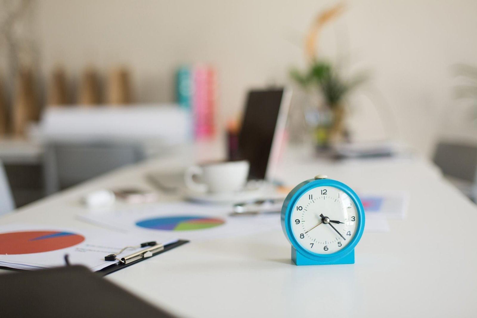 Um relógio de mesa azul-claro está em primeiro plano, sobre uma mesa branca de escritório. Ao fundo, fora de foco, há papéis com gráficos circulares, uma xícara de café, um laptop e outros objetos de trabalho. Como não há nenhuma pessoa à vista, o intuito da foto é retratar faltas e ausências no trabalho. 