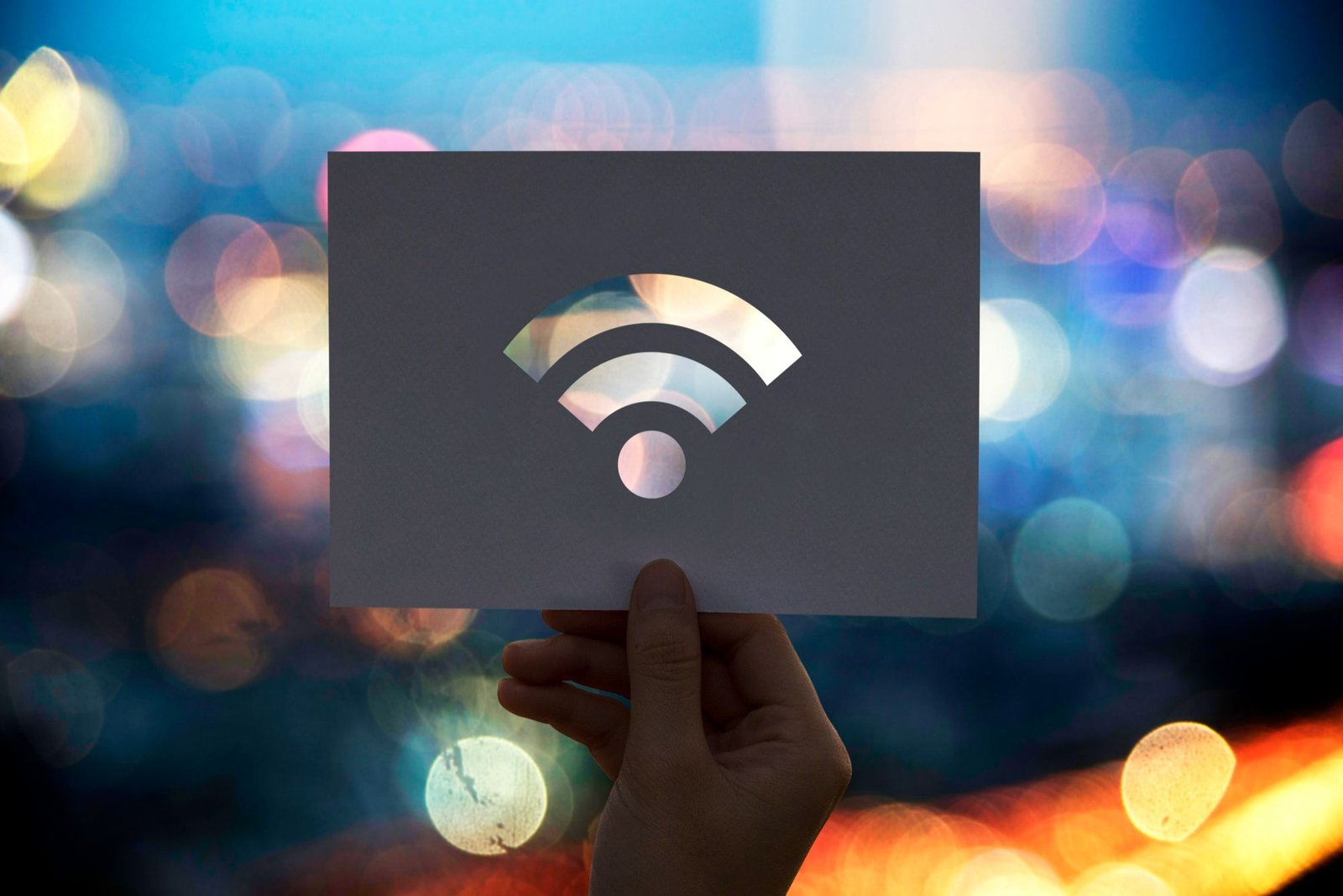 A foto mostra uma mão segurando um pedaço de papel cinza. No centro do papel, há um ícone de wi-fi recortado, revelando a luz brilhante de fundo. O fundo é desfocado, com luzes coloridas de uma cidade à noite, criando um efeito de bokeh.