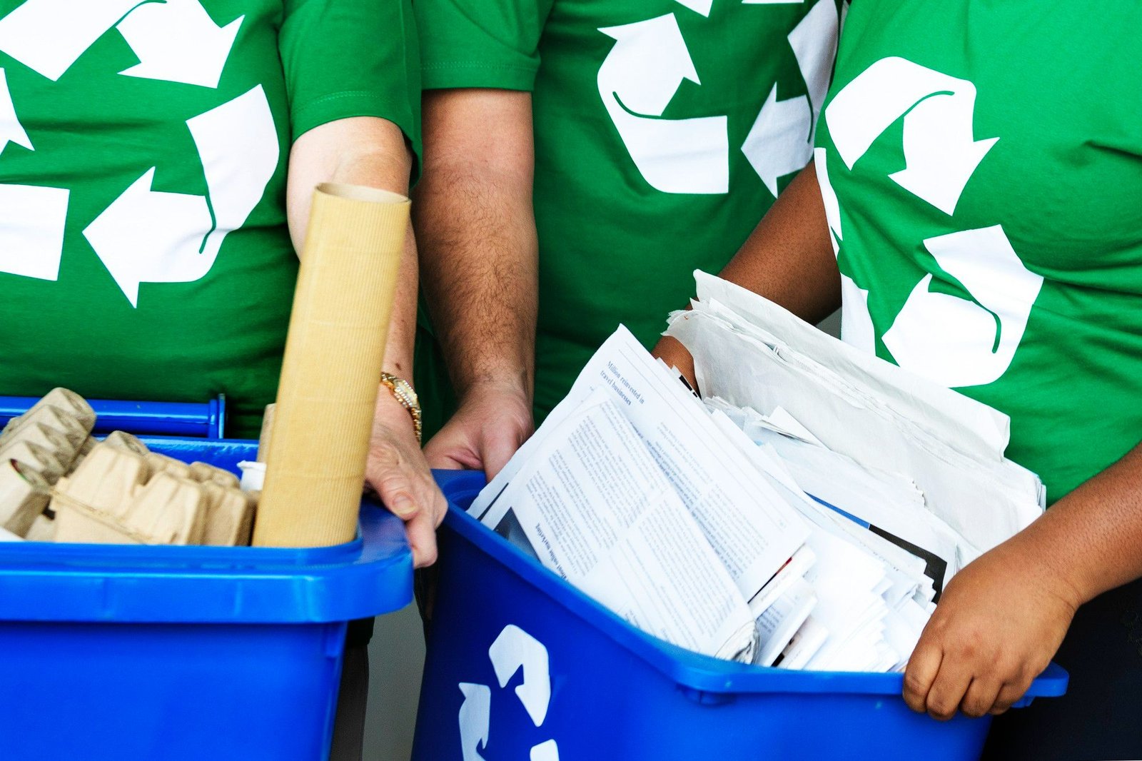 Três pessoas estão lado a lado, de frente, com a parte superior do corpo enquadrada. Todas usam camisetas verdes com o símbolo branco de reciclagem. Elas estão movendo duas caixas de reciclagem azuis. A caixa da esquerda contém papelão e rolos de papel; a da direita está cheia de papéis. A imagem visa ilustrar a importância do incentivo ao descarte correto do lixo em um hotel sustentável. 