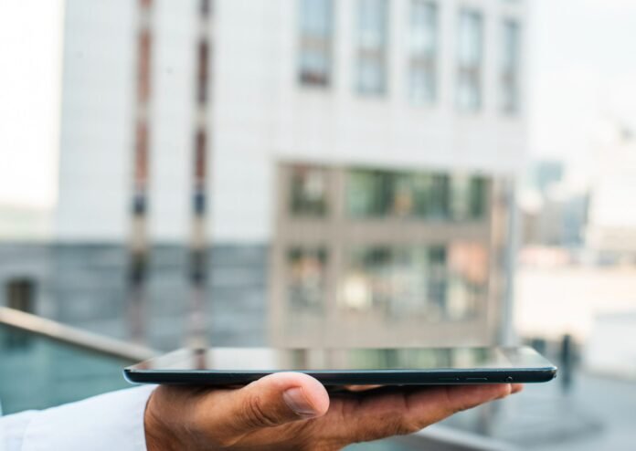 Homem de camisa social branca segura um tablet preto horizontalmente na palma da mão, com o polegar apoiado na lateral. O foco está na mão e no tablet, enquanto o fundo está desfocado, revelando a fachada de um prédio moderno com janelas grandes e uma grade de vidro. A foto simboliza a tecnologia aplicada à automação predial.