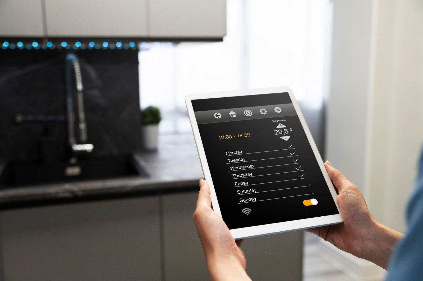 As mãos de uma pessoa seguram um tablet branco com uma interface de automação residencial visível na tela, mostrando a temperatura de 20.5ºC e a programação de horários para os dias da semana. A imagem, com o fundo desfocado de uma cozinha moderna (com pia e iluminação), ilustra como a tecnologia facilita o controle e a conveniência em ambientes como um quarto de hotel, aumentando a satisfação do hóspede e resultando em eficiência energética.