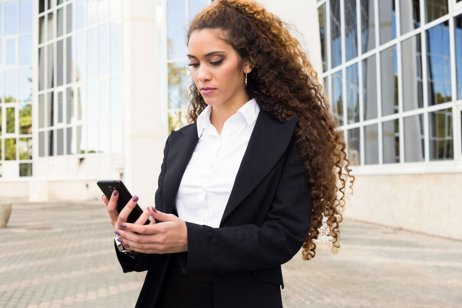 Uma mulher jovem, de cabelos cacheados longos e castanhos, vestindo um blazer preto e uma camisa social branca, está em pé em frente a um edifício moderno com janelas de vidro. Ela está concentrada em seu smartphone. A imagem simboliza a mobilidade e a conectividade, representando um gerente ou funcionário hoteleiro usando aplicativos para gerir tarefas ou acessar informações de qualquer lugar.