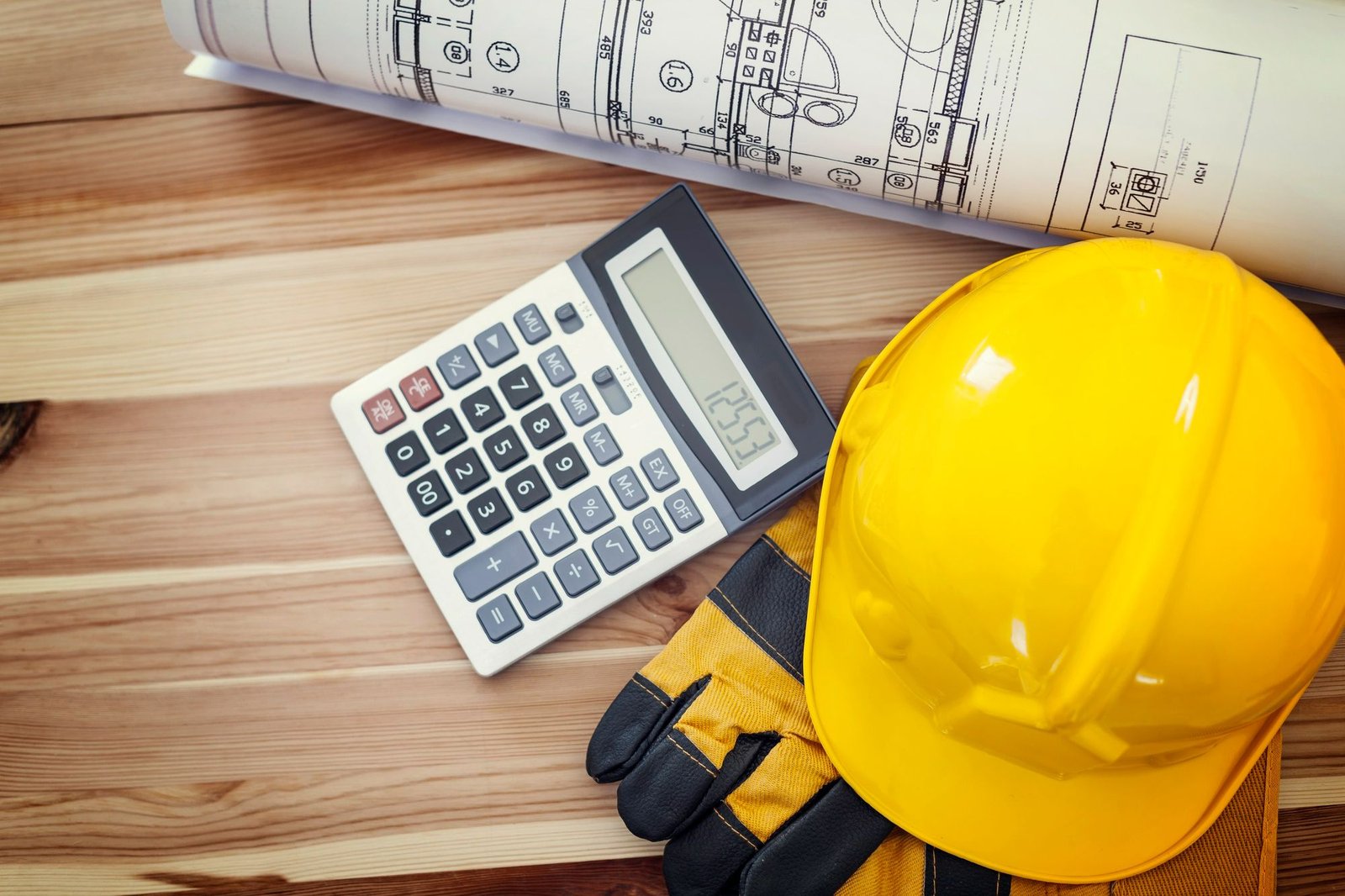 Vista superior de uma calculadora mostrando um número, junto a um capacete de segurança amarelo, luvas de proteção e plantas de projeto enroladas sobre uma mesa de madeira. A imagem representa o planejamento financeiro e o cálculo da economia de longo prazo gerada pela manutenção predial preventiva de hotéis.