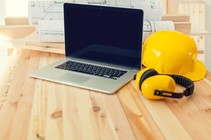 Imagem de um laptop aberto sobre uma mesa de trabalho de madeira clara, com projetos ou plantas de fundo e, ao lado, um capacete de segurança amarelo e protetores auriculares. A imagem ilustra o planejamento e a gestão da manutenção predial em hotéis.