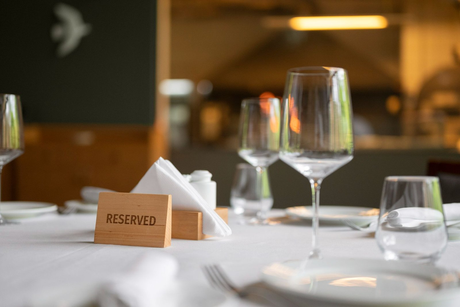 Mesa de restaurante formal e elegantemente posta com talheres, pratos brancos, taças de vinho e um guardanapo branco. Em primeiro plano, uma pequena placa de madeira diz "RESERVED" (reservado). O fundo do restaurante está sutilmente iluminado. A foto representa o serviço personalizado oferecido pela recepção do hotel, que cuida de reservas e outros arranjos, garantindo uma experiência impecável e sem preocupações.
