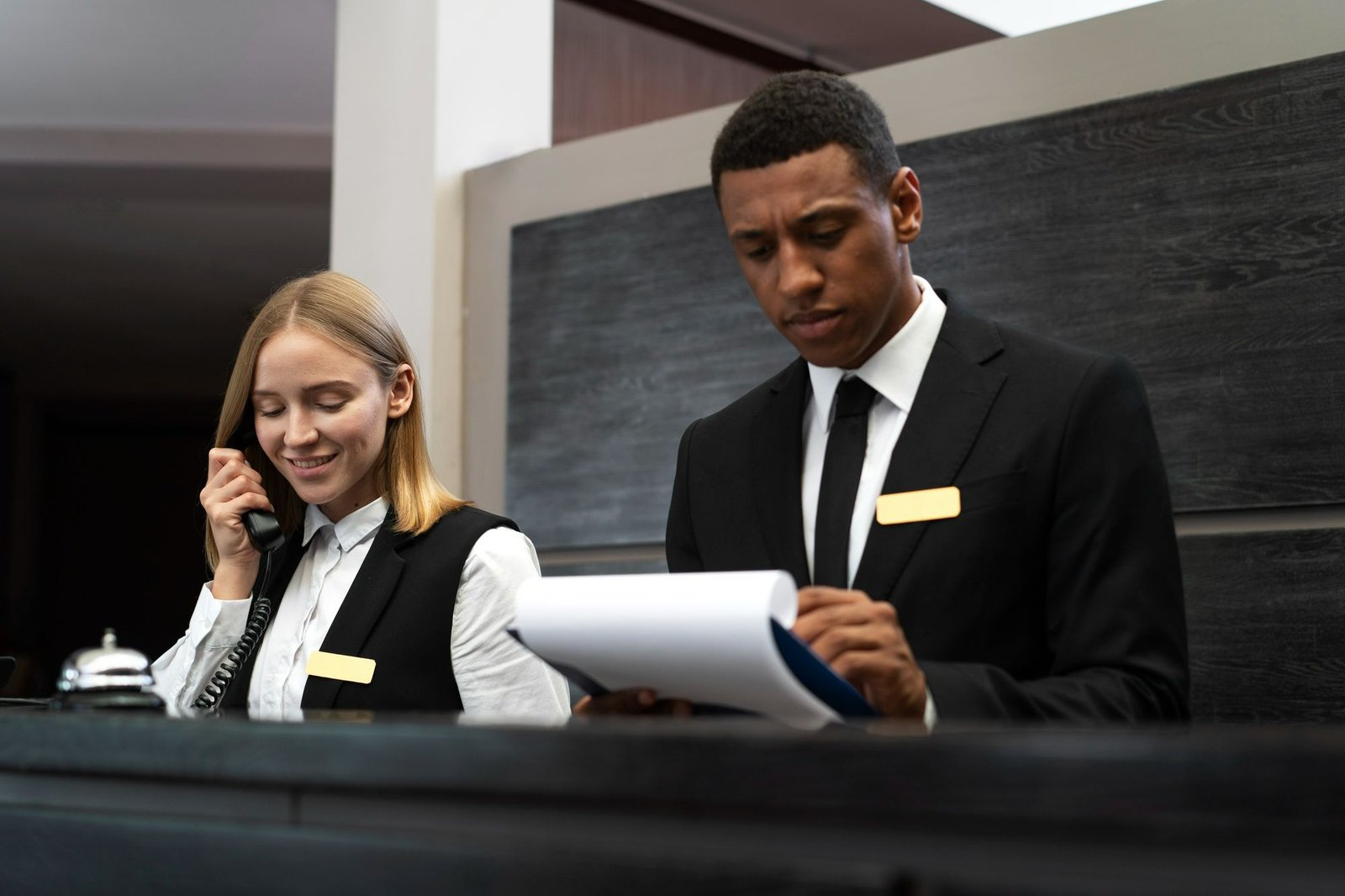 Dois recepcionistas uniformizados (um homem e uma mulher) trabalham atrás de um balcão moderno. A recepcionista está sorrindo enquanto atende a um telefone, e o recepcionista ao lado examina cuidadosamente uma prancheta com documentos. A imagem simboliza a necessidade de a recepção possuir um banco de dados atualizado com prestadores de serviço e dicas do destino para oferecer recomendações sob medida.
