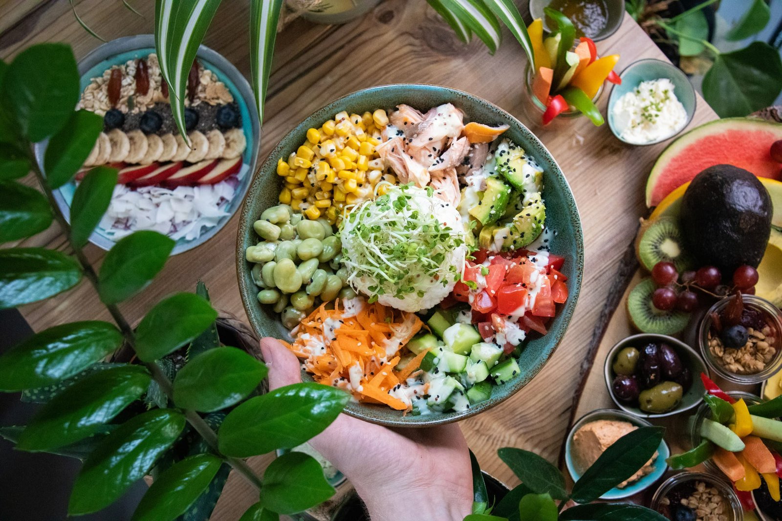 Uma vista aérea em close-up de uma mão segurando uma grande tigela de 'poke' ou 'buddha bowl' colorida, repleta de ingredientes frescos. A tigela contém, em seções separadas, milho doce, fatias de salmão (ou atum, se for um 'poke' clássico), abacate picado, tomate picado, pepino, cenoura ralada com molho, favas verdes e um montinho central de brotos. Ao redor, em uma mesa de madeira, há outras opções de alimentos saudáveis e frescos, como um 'smoothie bowl' de frutas e granola, e uma tábua com melancia, kiwi, abacate inteiro, uvas, azeitonas, pimentões, e diferentes patês ou molhos. A foto pretende ilustrar que o hotel dining se fortalece quando o restaurante se dispõe a ter um cardápio diversificado.