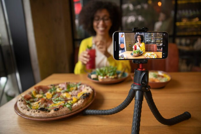 Uma mulher sorridente, com cabelo encaracolado e óculos, está sentada à mesa de um restaurante, segurando um copo com uma bebida vermelha. Em frente a ela há uma pizza gourmet e outros pratos. Um smartphone em um tripé flexível está posicionado na mesa, gravando a cena. A tela do celular mostra a mulher e a comida, indicando que ela está criando conteúdo, como um vídeo ou transmissão ao vivo, sobre sua experiência gastronômica.