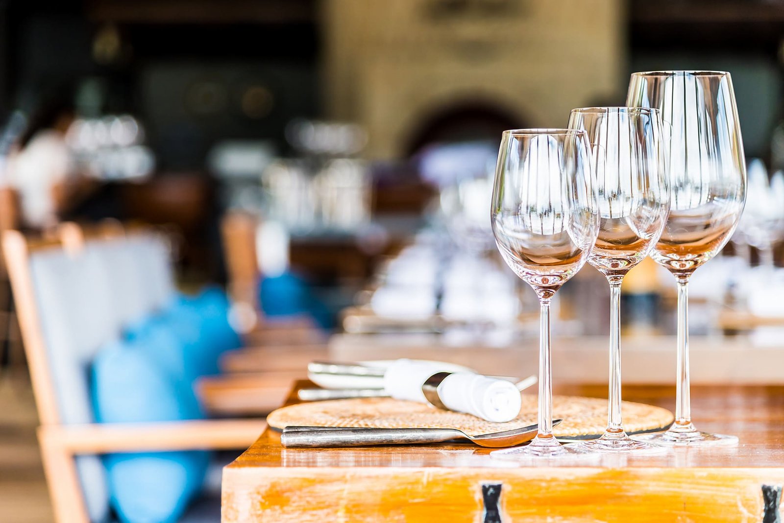Três taças de vinho vazias estão dispostas em uma mesa de madeira polida, em um ambiente similar ao de restaurantes de hotéis. Ao lado das taças há um descanso de mesa individual com um guardanapo branco enrolado e talheres. O fundo está desfocado, mas sugere um terraço ou área externa sofisticada, com uma cadeira de madeira e almofadas azuis visíveis à esquerda. A foto evoca uma atmosfera de refeição de luxo e relaxamento.