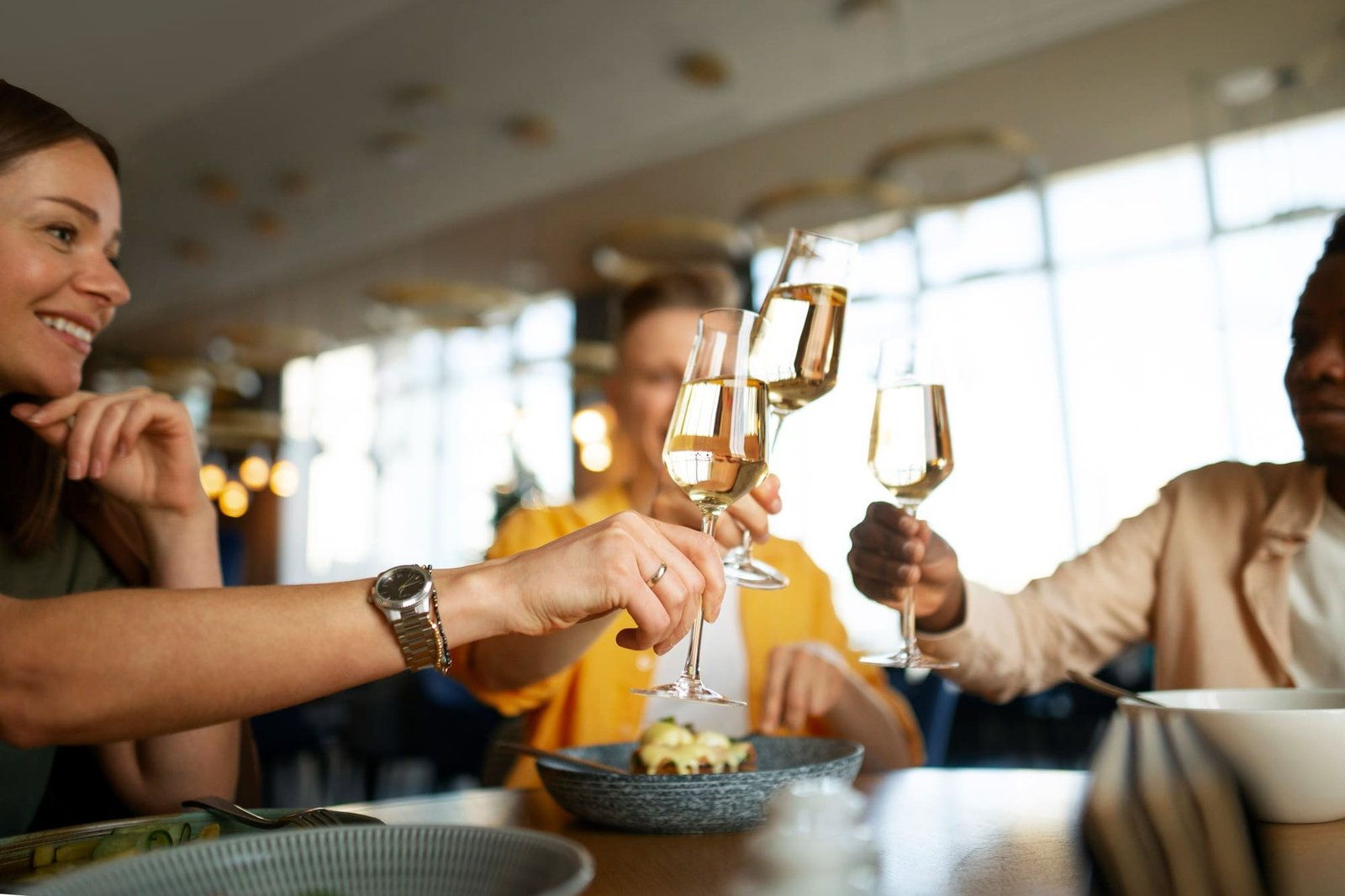 Quatro pessoas, sorrindo, estão fazendo um brinde com taças de vinho branco em um restaurante de hotel elegante e bem iluminado. A foto está focada nas taças e nas mãos que se tocam no brinde, com uma mulher sorridente em primeiro plano à esquerda. A mulher está usando um relógio de pulso e um anel. Em primeiro plano, sobre a mesa, há pratos com comida. O fundo mostra as luminárias modernas e o ambiente sofisticado do local.