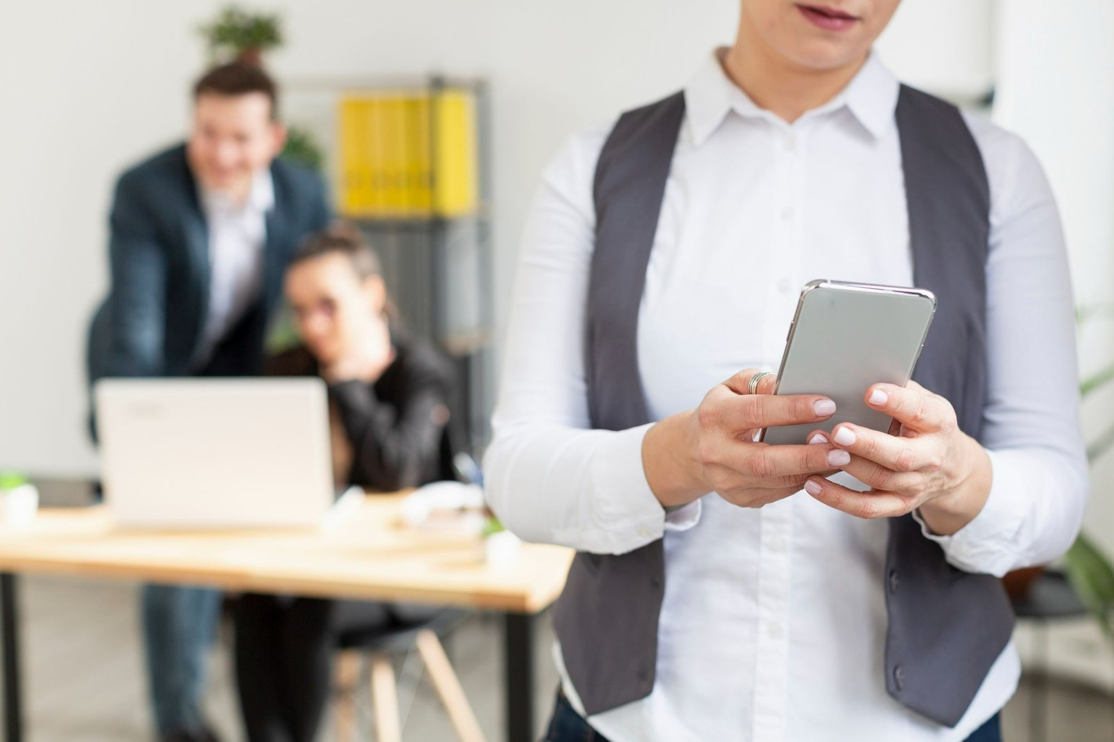 Uma mulher, vestindo uma camisa social branca e um colete cinza, utiliza seu smartphone, representando o uso de tecnologia (como o WhatsApp) pelo RH hoteleiro. Em segundo plano e fora de foco, um homem e uma mulher interagem em frente a um computador numa mesa de escritório.