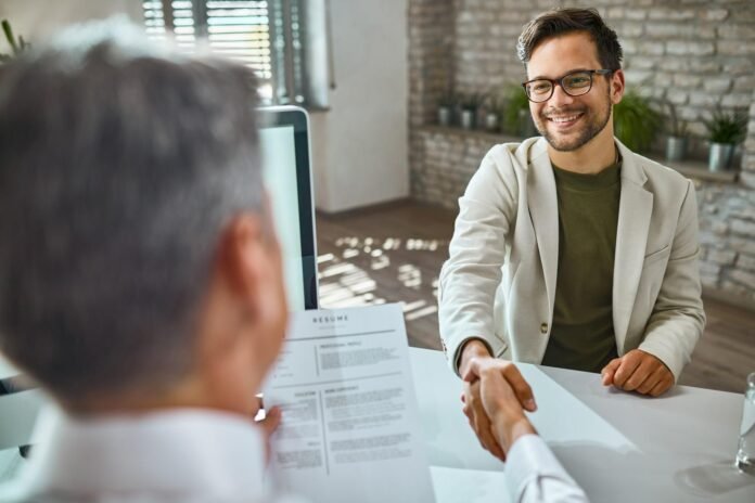 Jovem candidato masculino, sorridente e de óculos, vestindo um blazer claro sobre uma camisa verde-escura, aperta a mão de um recrutador durante uma entrevista de emprego. O candidato está sentado à frente de uma mesa clara, e na mão do recrutador está parcialmente visível um currículo. A cena simboliza o recrutamento hoteleiro.