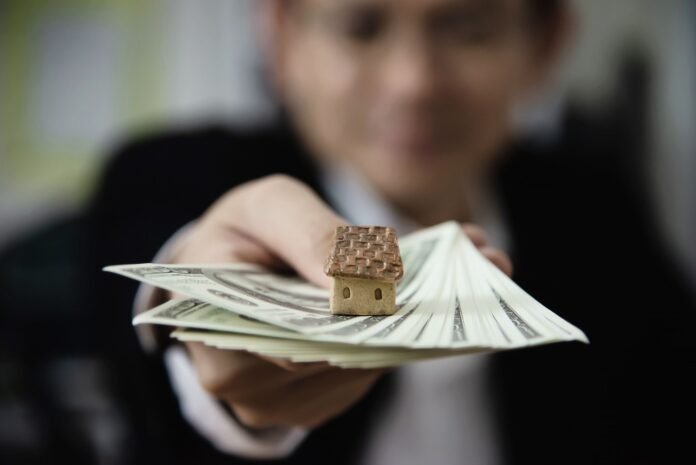9 - Marketing & Vendas - Linhas de crédito para hotéis - Crédito - Freepik - 2 Uma mão estendida em direção à câmera segura diversas notas de dólar, sobre as quais está uma casa em miniatura com telhado marrom. Ao fundo, uma pessoa de terno escuro está desfocada. A imagem pretende ilustrar a adesão a linhas de crédito para hotéis.