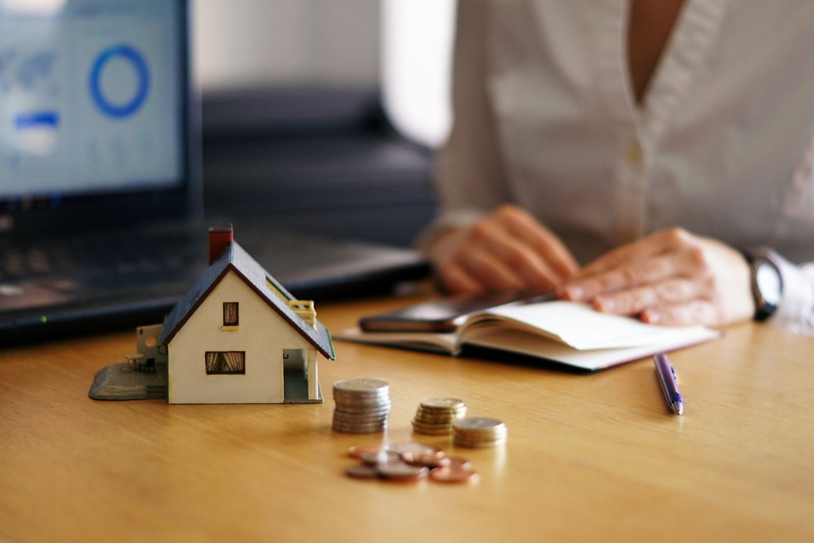 Mesa de escritório com uma casa em miniatura, moedas empilhadas em primeiro plano e, ao fundo, uma pessoa analisando documentos em um caderno aberto, com um notebook exibindo gráficos de análise financeira. A imagem representa a consulta e o processo de análise de crédito por instituições financeiras para o setor hoteleiro.