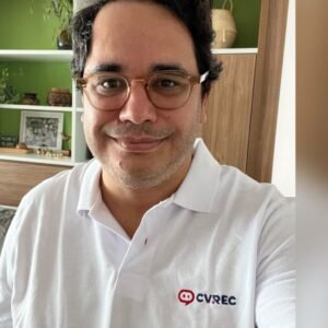 Foto selfie de Rodrigo Garcez, homem de pele clara e cabelo escuro, usando óculos de armação clara e camisa polo branca com a logomarca vermelha e preta "CVREC". Ele sorri gentilmente para a câmera. O fundo desfocado é um ambiente interno com prateleiras e objetos de decoração.