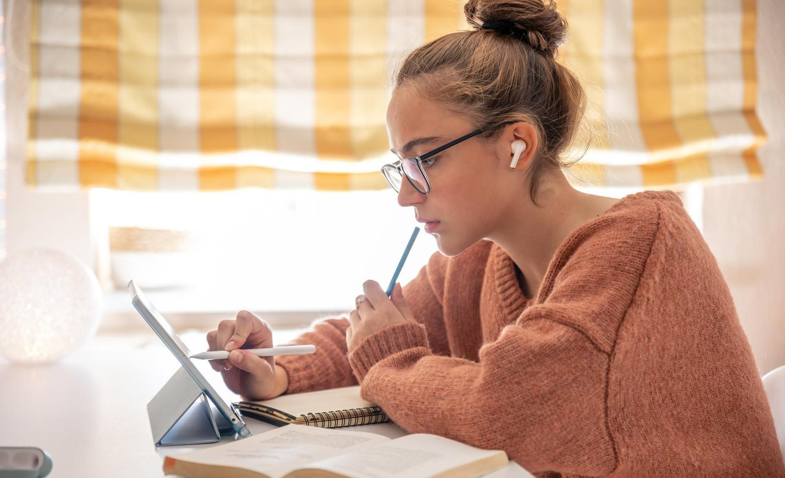 Jovem com óculos e coque, vestindo um suéter salmão, usa fones de ouvido sem fio enquanto trabalha concentrada em um tablet apoiado em uma mesa. Ela segura uma caneta digital e está cercada por livros e cadernos. A imagem sugere estudo, aprendizado online ou pesquisa.