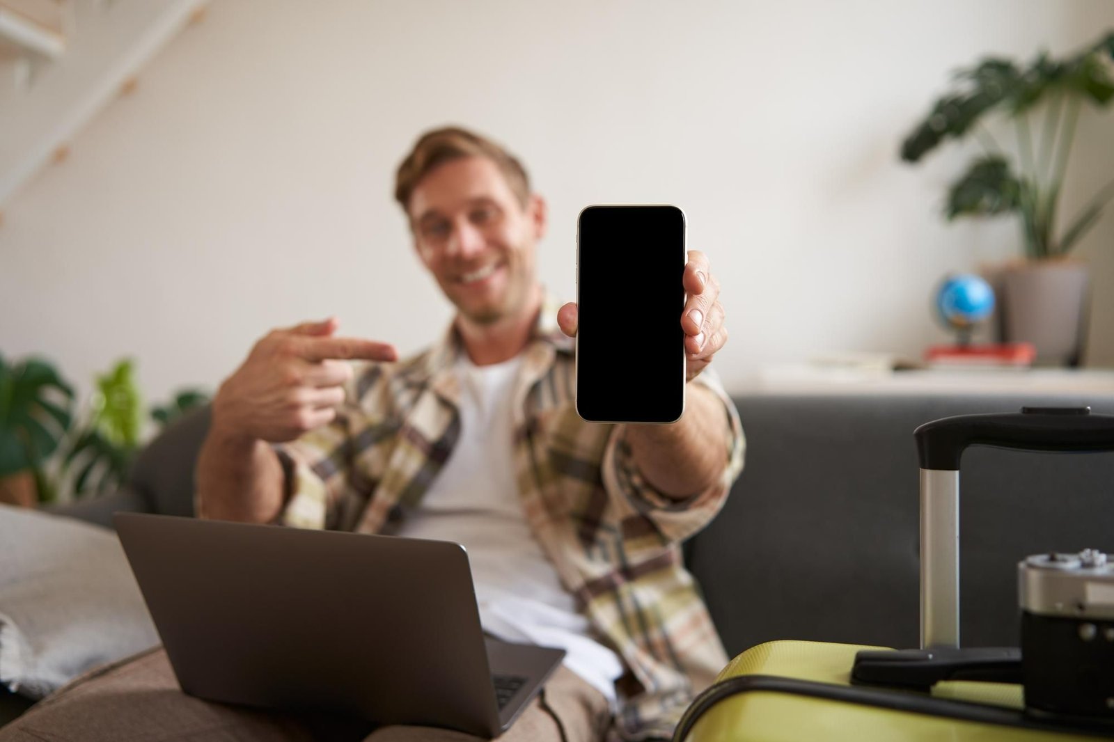 Um homem jovem e sorridente, sentado em um sofá com um laptop no colo, aponta o dedo indicador para a tela escura e em branco de um smartphone que ele segura na frente. A imagem foca no celular, enquanto, desfocadas, estão uma mala amarela e uma câmera de fotos ao seu lado direito.