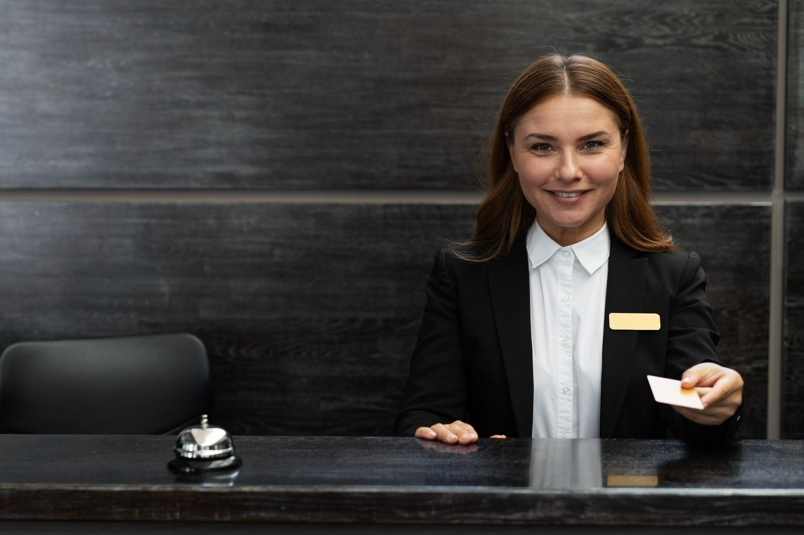 Uma recepcionista de hotel sorridente, vestindo um blazer preto e camisa branca com um crachá dourado, estende a mão para entregar um cartão-chave (key card) no balcão de check-in. O balcão é de madeira escura e, à esquerda, há um pequeno sino de recepção prateado. O fundo é um painel de madeira escura com ranhuras horizontais.