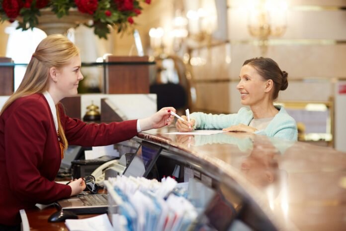 Uma recepcionista de hotel sorridente, vestindo um blazer vermelho, entrega uma caneta a uma hóspede madura, também sorridente, que se apoia no balcão da recepção para preencher um documento. O ambiente é elegante, com iluminação quente, e reflete o atendimento personalizado em hotéis.
