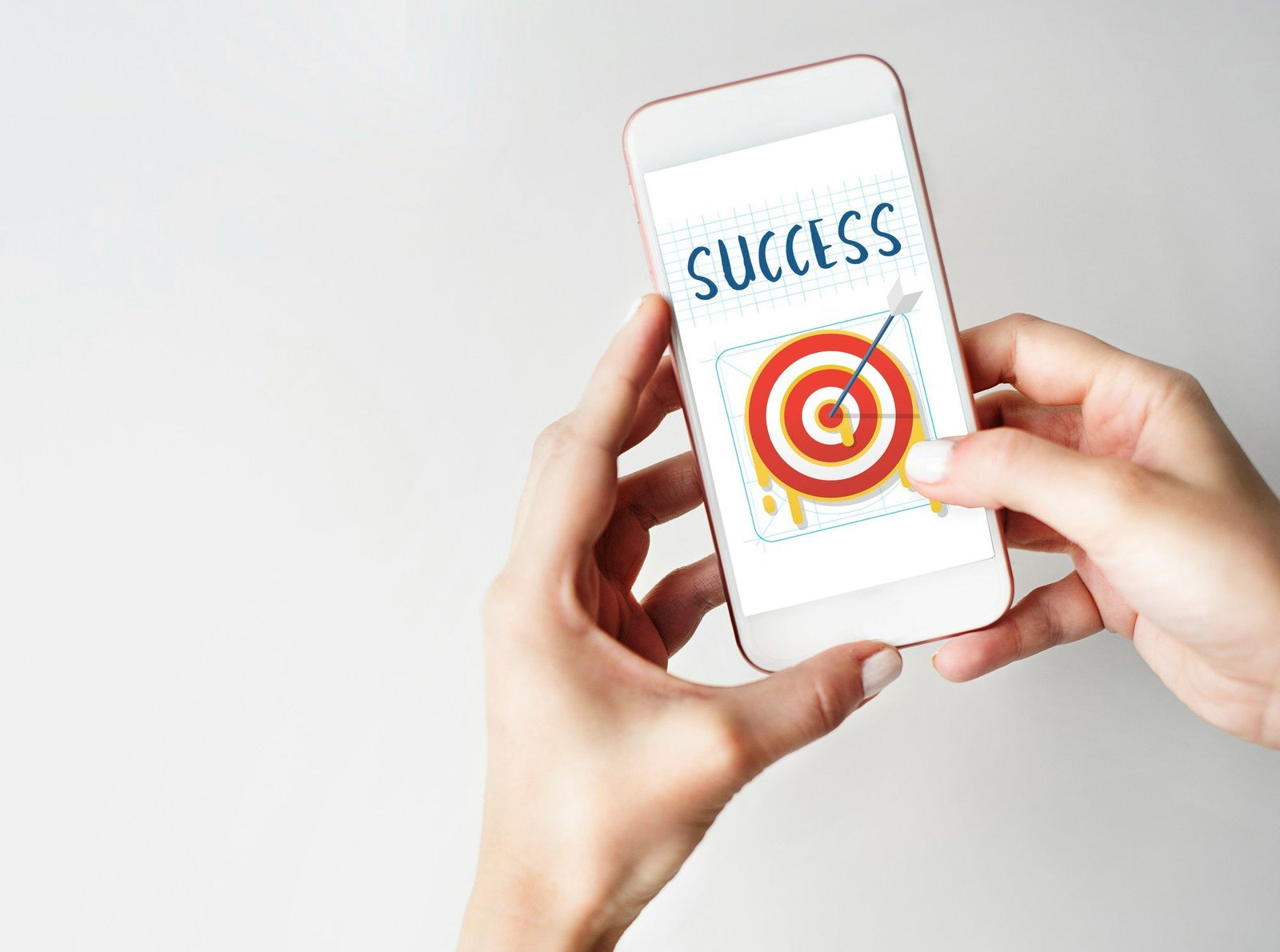Close-up de mãos segurando um smartphone branco com a palavra 'SUCCESS' (SUCESSO) exibida na tela. Abaixo do texto há um gráfico de alvo concêntrico (target) com uma flecha perfeitamente cravada no centro, simbolizando o alcance de metas e o foco no resultado, essenciais para uma estratégia de marketing digital eficaz.