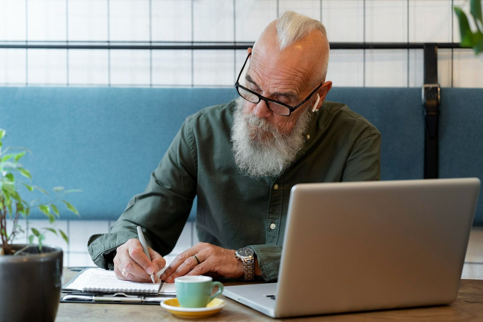 Um homem mais velho, com barba e cabelo branco característicos de um baby boomer, escreve em um caderno e parece trabalhar concentrado. Ele usa óculos e fones de ouvido sem fio. Ao lado do caderno estão um notebook aberto e uma xícara de café. A imagem visa representar a importância de a gestão de pessoas na hotelaria não se guiar única e exclusivamente pela idade ao contratar para vagas de emprego. 