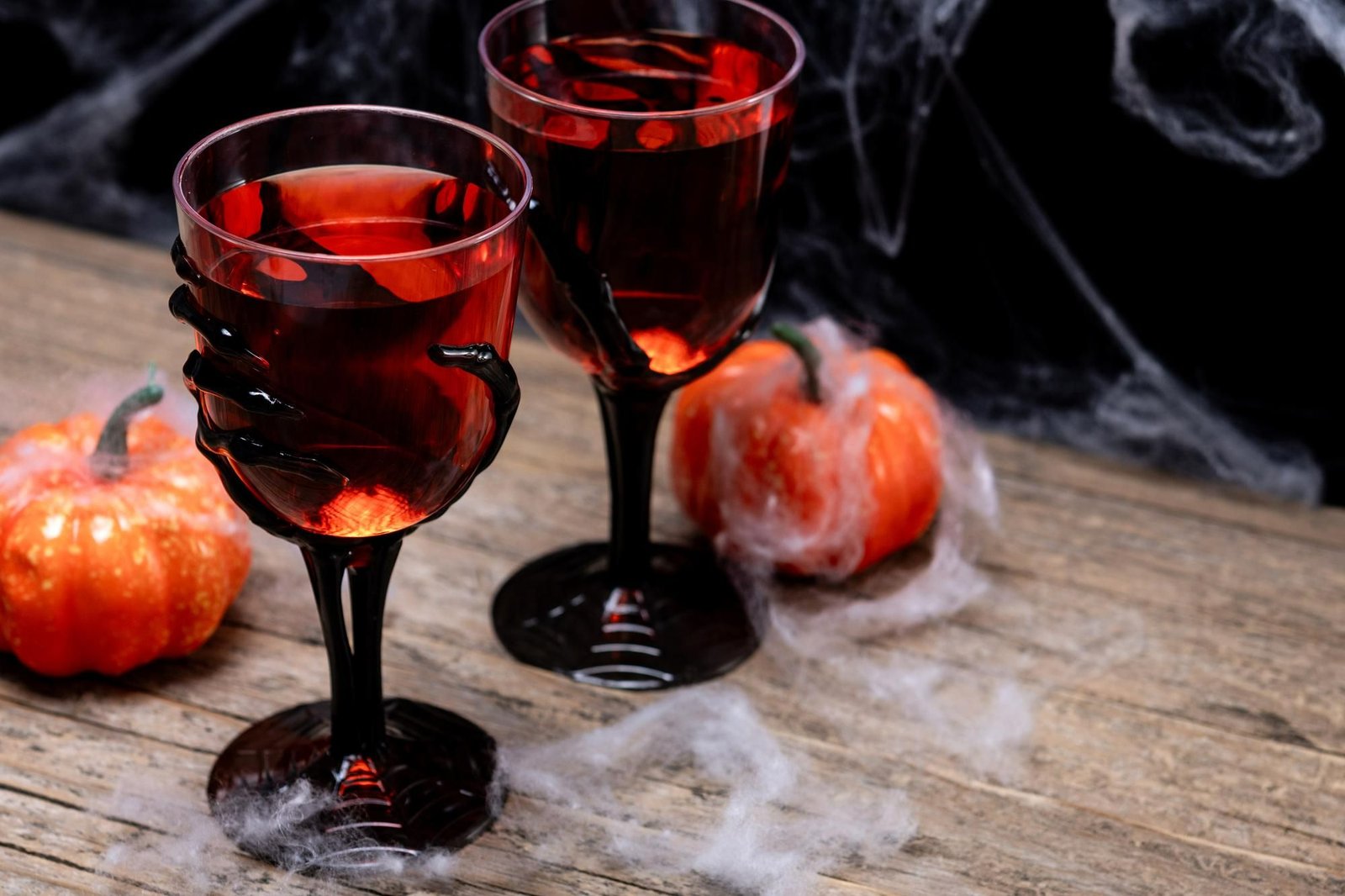 Dois cálices de Halloween pretos com líquido vermelho sobre uma mesa de madeira rústica, decorados com abóboras em miniatura e teias de aranha.