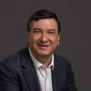 Retrato em close-up de Jeferson Munhoz, um homem de pele morena clara e cabelos escuros, levemente grisalhos nas laterais, sorrindo. Ele veste um blazer azul-marinho sobre uma camisa social branca de colarinho aberto. O fundo é de cor cinza-escuro sólido.