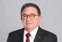 ABIH Nacional: 89 anos fortalecendo a hotelaria e impulsionando o turismo brasileiro Retrato em estúdio de Manoel Cardoso Linhares, presidente da ABIH Nacional. Ele é um homem maduro, de pele clara, usando óculos de aro marrom, terno preto, camisa social branca e uma gravata bordô com padrão xadrez. Ele está olhando diretamente para a câmera, com uma expressão neutra e profissional. Um pequeno broche prateado está fixado na lapela direita do terno. O fundo é de cor cinza-claro e uniforme.