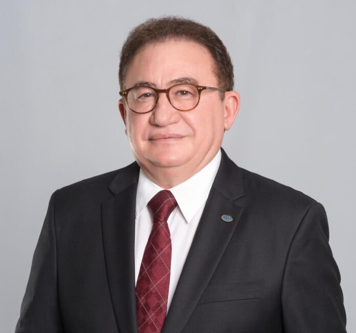 Retrato em estúdio de Manoel Cardoso Linhares, presidente da ABIH Nacional. Ele é um homem maduro, de pele clara, usando óculos de aro marrom, terno preto, camisa social branca e uma gravata bordô com padrão xadrez. Ele está olhando diretamente para a câmera, com uma expressão neutra e profissional. Um pequeno broche prateado está fixado na lapela direita do terno. O fundo é de cor cinza-claro e uniforme.