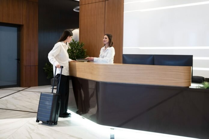 Uma recepcionista de hotel sorridente, vestindo uma camisa social branca, atende uma hóspede em uma recepção moderna com acabamento em madeira. A cliente, também de camisa branca e com uma calça escura à mostra, está ao lado do balcão segurando a alça de uma mala de viagem. A cena destaca o atendimento pessoal e acolhedor na chegada ao hotel.