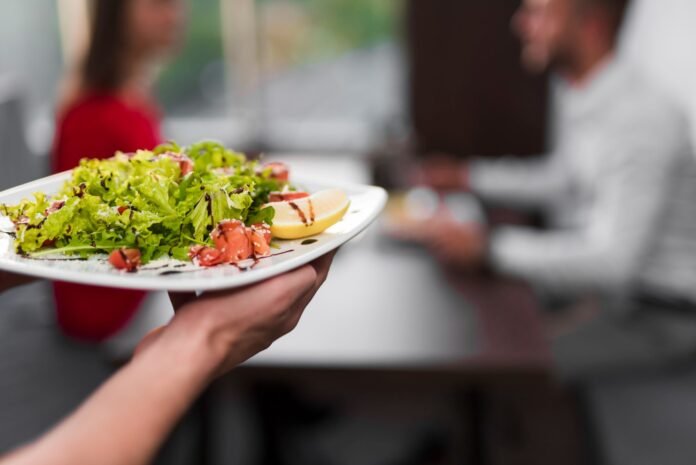 Prato de salada com apresentação moderna e ingredientes frescos sendo servido em um restaurante. A imagem destaca uma refeição saudável e nutritiva, uma das principais tendências em Alimentos e Bebidas. No fundo desfocado, dois clientes estão sentados à mesa e conversando.