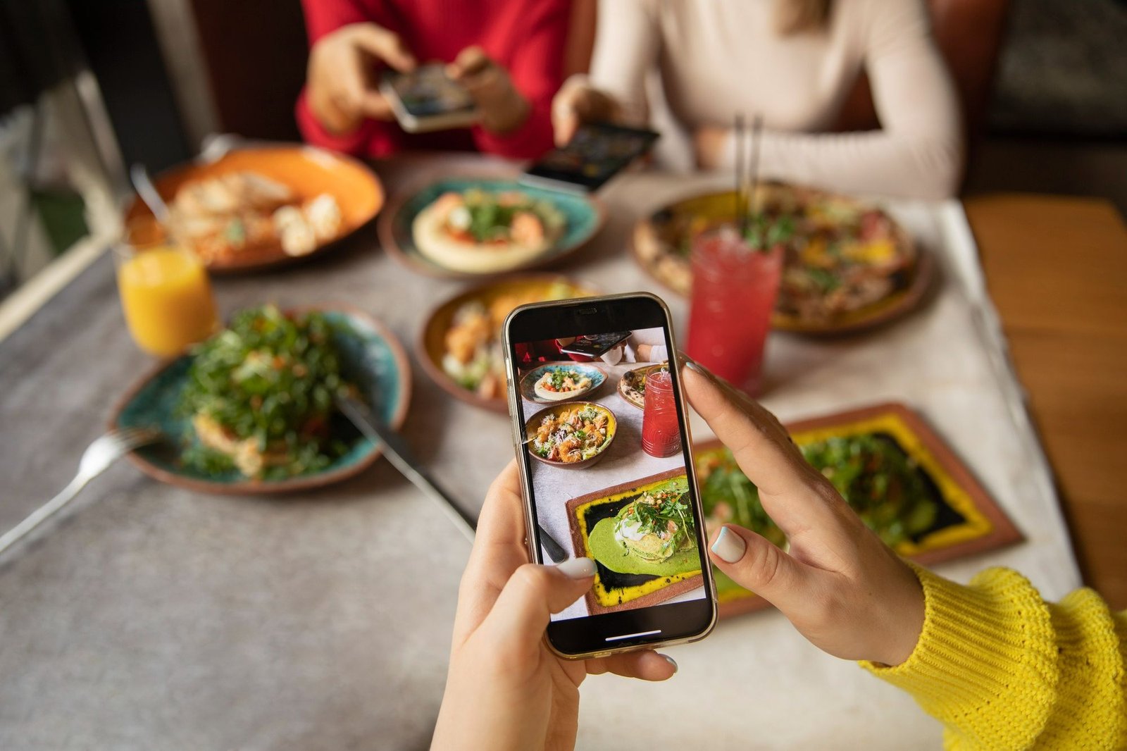 Pessoas em um restaurante utilizando smartphones para fotografar pratos gourmet e bebidas coloridas. A imagem ilustra a tendência de pratos 'instagramáveis' e a digitalização da experiência gastronômica, na qual o compartilhamento visual é parte essencial da refeição.