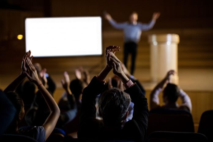 Pessoas aplaudindo em uma plateia escura, com as mãos levantadas, enquanto um palestrante está no palco, em frente a uma tela branca iluminada, com os braços abertos em um gesto de agradecimento. O foco está nas mãos da plateia.
