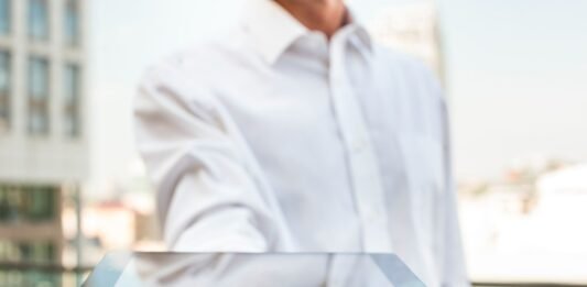 Um homem, vestindo uma camisa social branca, estende um tablet preto em primeiro plano, como se o estivesse oferecendo ou então mostrando a tela. O tablet sugere o uso de tecnologia para serviços como check-in online. Ao fundo, fora de foco, vê-se um prédio moderno em um dia claro, reforçando a ideia de retrofit tecnológico hoteleiro.