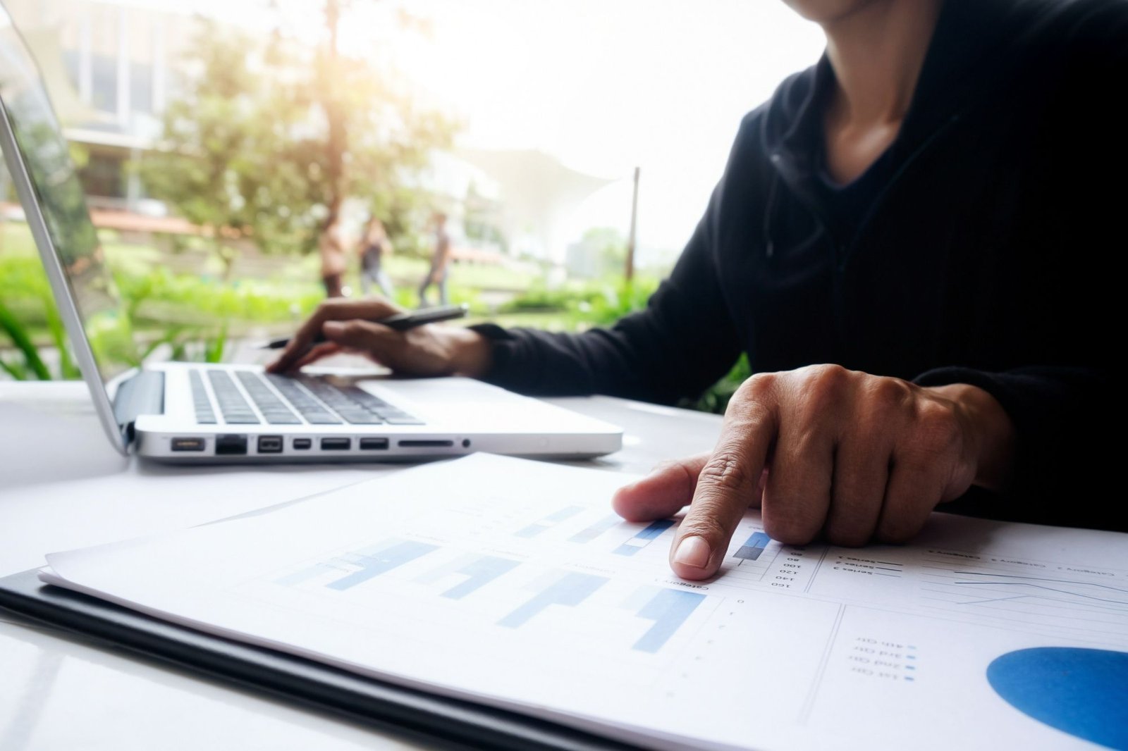 Close-up de uma pessoa trabalhando em uma mesa branca. A pessoa, vestindo uma blusa preta, usa a mão direita para digitar em um notebook prateado, enquanto a mão esquerda aponta para um gráfico de barras impresso em uma folha sobre a mesa, indicando análise de dados ou planejamento.