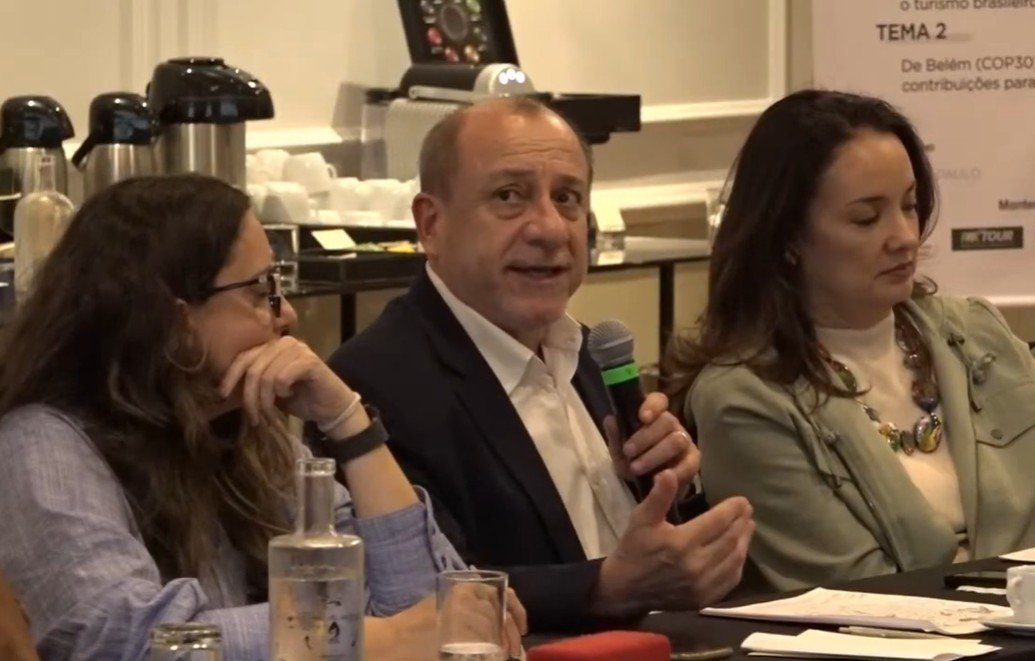 Toni Sando fala ao microfone, sentado entre Marina Figueiredo e Carolina Negri em uma mesa de debate durante o evento.