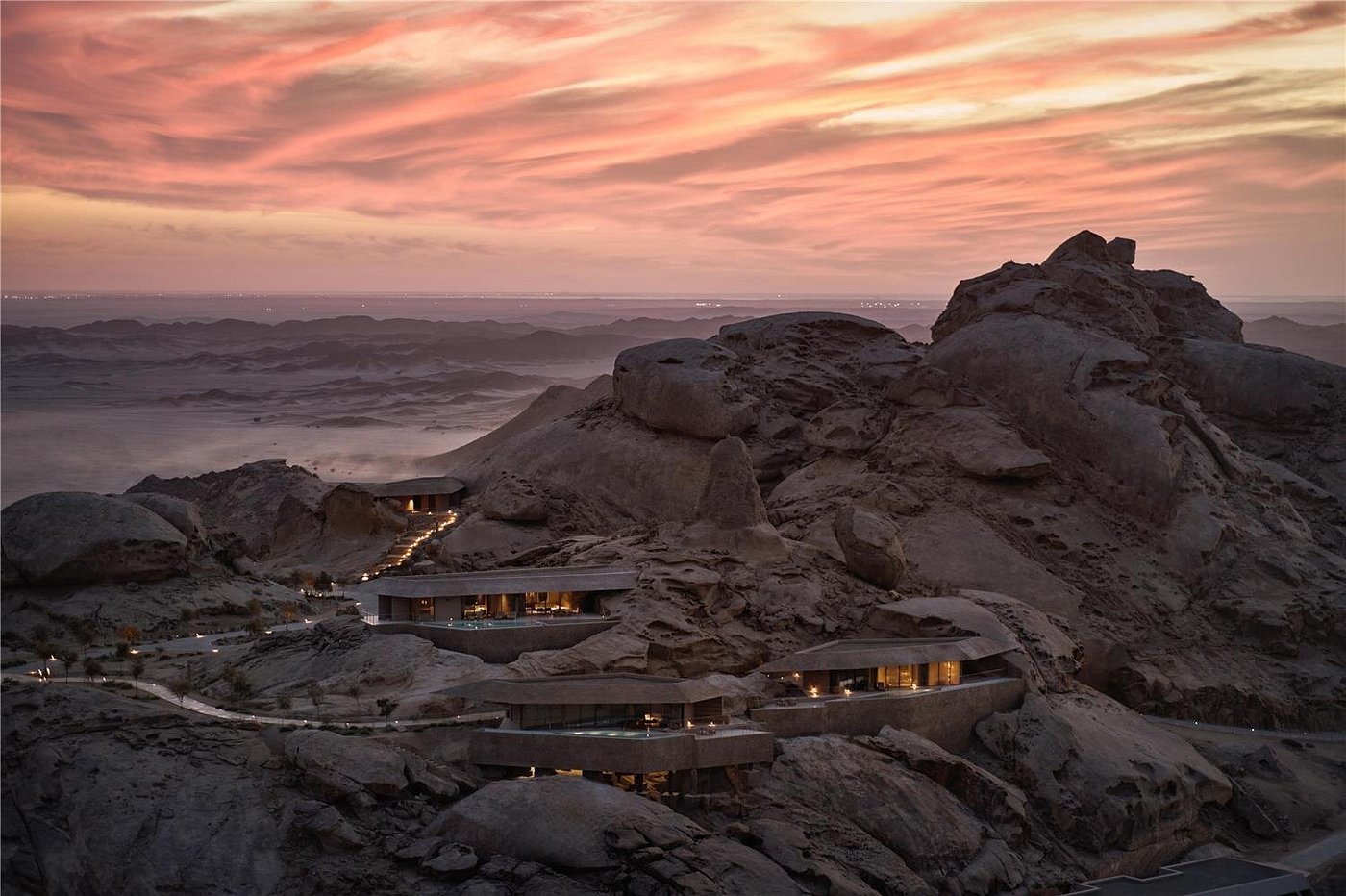 Vista panorâmica do Desert Rock Resort ao entardecer. As estruturas arquitetônicas modernas e luxuosas estão integradas e esculpidas diretamente nas encostas de montanhas rochosas em um deserto. O céu está tingido com tons vibrantes de rosa, laranja e roxo, enquanto as luzes internas das suítes brilham suavemente, criando um contraste acolhedor com a pedra bruta.