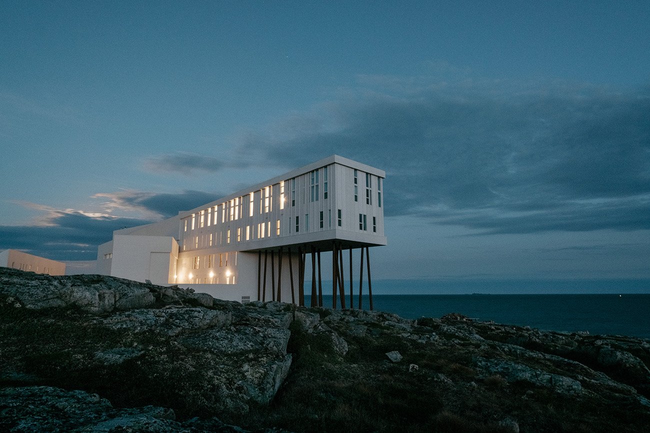Fotografia ao entardecer do hotel Fogo Island Inn, no Canadá. O edifício branco, com arquitetura moderna e minimalista, destaca-se em uma costa rochosa e escura. Uma extremidade do prédio é suspensa por estacas altas, projetando-se em direção ao mar. As janelas estão iluminadas, criando um brilho acolhedor contra o céu azul profundo e nublado.