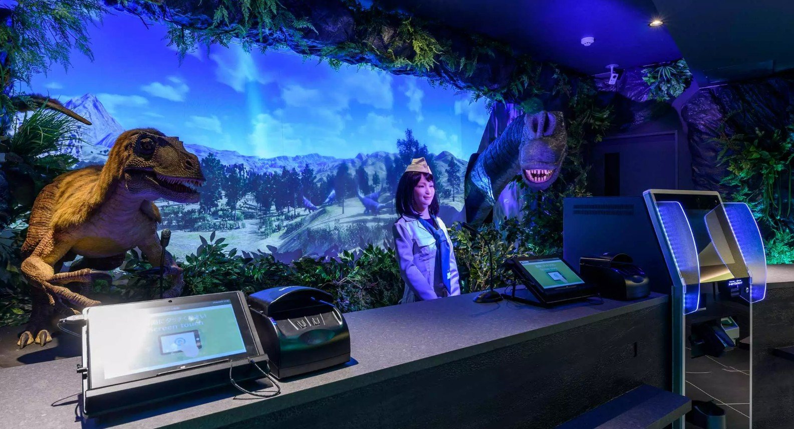 Foto da recepção do Henn na Hotel Tokyo Haneda. Atrás de um balcão moderno com telas de autoatendimento estão três robôs: uma atendente humanoide ao centro e dois dinossauros (um raptor à esquerda e um de pescoço longo à direita). O ambiente é decorado com folhagens e iluminado por luzes azuis, com um painel ao fundo exibindo uma paisagem pré-histórica.