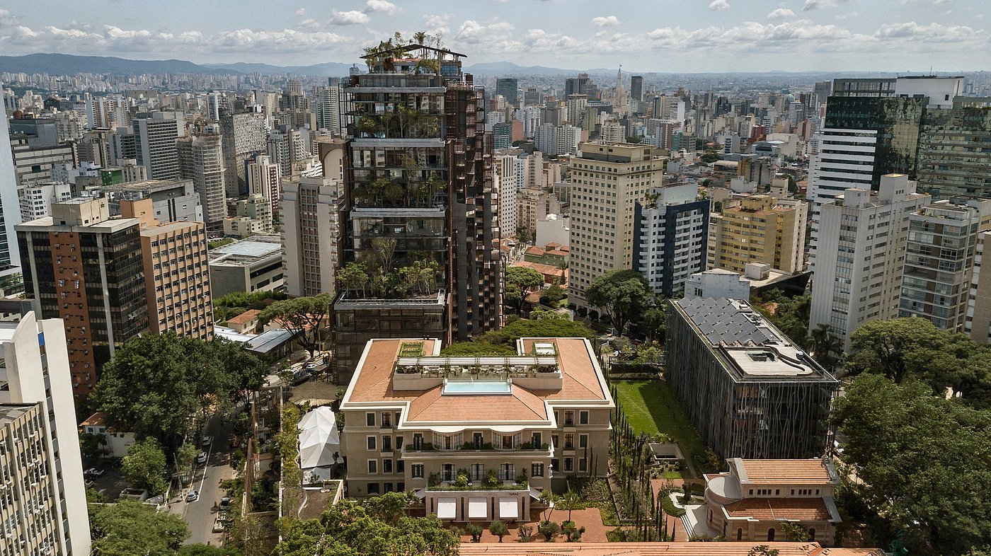 Vista aérea do complexo Rosewood São Paulo. Em destaque, a "Torre Mata Atlântica", um edifício alto coberto por vegetação exuberante, ao lado de um prédio histórico restaurado com uma piscina no topo. O hotel está inserido em uma densa paisagem urbana, cercado pelos arranha-céus e ruas arborizadas da capital paulista sob um céu claro.