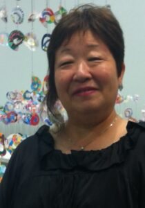 Retrato de rosto de Elza Tsumori, a autora deste artigo sobre marketing de experiência. Ela é uma mulher de ascendência oriental, com cabelos curtos e escuros levemente ondulados, e sorri suavemente para a câmera. Veste uma blusa preta com decote franzido e usa um colar delicado. Ao fundo, decorações coloridas em formato circular aparecem desfocadas.