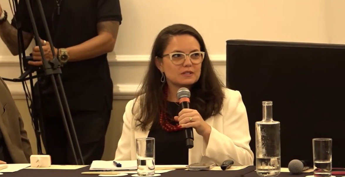 Jaqueline Gil, usando óculos, blazer branco e um colar vermelho, fala em um microfone ao conduzir o evento que promoveu o debate acerca das mudanças climáticas e seus impactos no turismo. À sua frente estão uma garrafa d'água e copos; ao fundo, um operador de câmera com equipamento.