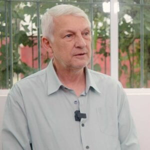 Retrato em plano médio de José Roberto Muratori, um homem de cabelos grisalhos e curtos, vestindo uma camisa social verde-clara. Ele está posicionado em um ambiente interno, olhando ligeiramente para a direita da câmera. Um pequeno microfone de lapela está preso à sua camisa. O fundo mostra uma janela com grades e vegetação.