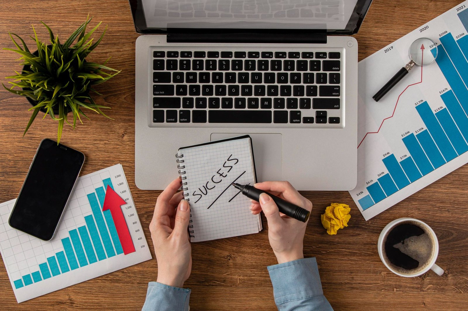 Vista superior de uma mesa de madeira com um notebook, smartphone, xícara de café e gráficos de barras que mostram crescimento com setas vermelhas para cima. No centro, mãos seguram uma caneta e um bloco de notas no qual está escrita a palavra "SUCCESS" (sucesso).