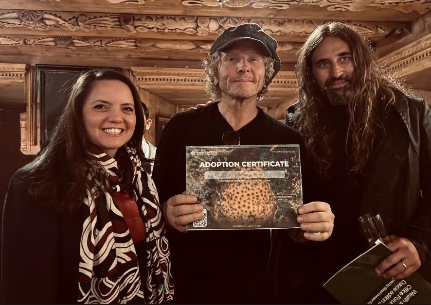 Fotografia mostra Jaqueline Gil, Matt Sorum e Pablo Lobo em um ambiente interno com teto e paredes de madeira entalhada em Davos. À esquerda, Jaqueline Gil sorri usando um casaco escuro e um lenço estampado em tons de vinho e branco. No centro, o músico e investidor Matt Sorum, vestindo uma camiseta preta e boina cinza, segura um "Adoption Certificate" (Certificado de Adoção) da Biofábrica de Corais, que exibe a foto de um coral em regeneração. À direita, Pablo Lobo, com cabelos longos e casaco escuro, segura alguns folhetos do evento enquanto posa para a foto. O registro captura o momento da entrega do certificado, simbolizando o apoio a projetos socioambientais na costa brasileira.