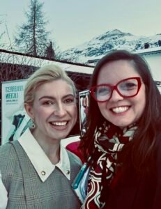 Fotografia em estilo selfie de Anastasia Kalinina (à esquerda) e Jaqueline Gil (à direita), sorrindo em um ambiente externo em Davos. Anastasia tem cabelos loiros presos e usa um colete xadrez cinza sobre uma camisa branca com botões dourados na gola. Jaqueline Gil usa óculos de armação vermelha e um lenço estampado em tons de preto, branco e vermelho. Ao fundo, vê-se um cenário invernal com uma montanha coberta de neve sob um céu claro, um pinheiro alto e parte de um banner do evento. A imagem registra a parceria entre as duas especialistas durante o encontro do Basel Investor Forum 2026.