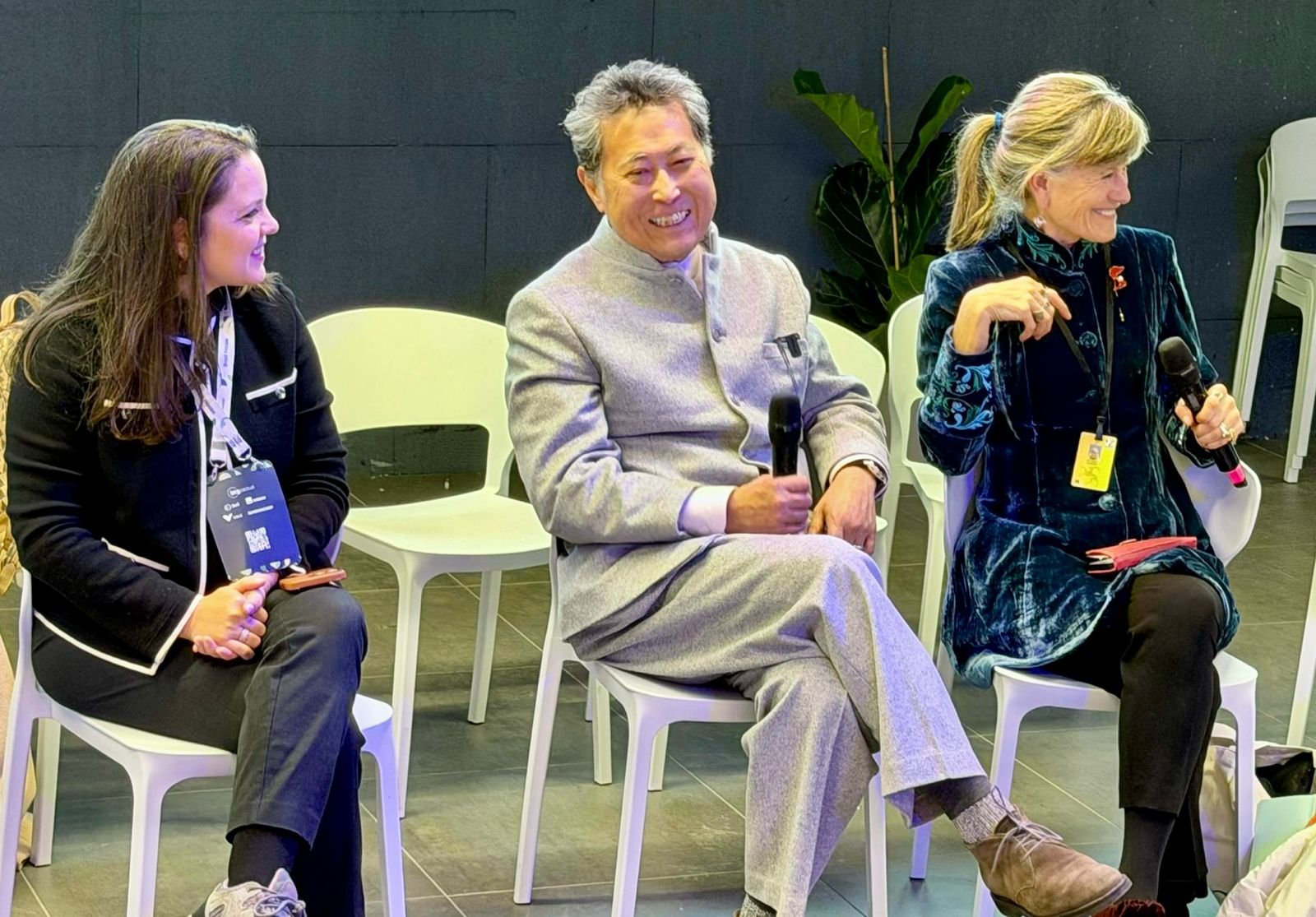 Fotografia de um painel de discussão em Davos, apresentando três lideranças sentadas em cadeiras brancas modernas. À esquerda, Jaqueline Gil sorri, vestindo um blazer preto com detalhes em branco e usando um crachá de identificação. No centro, Chavalit Frederik Tsao ri de forma descontraída, vestindo um terno cinza de gola alta e segurando um microfone. À direita, Jacqueline Novogratz também sorri enquanto segura um microfone, vestindo um casaco de veludo azul-turquesa com bordados delicados. O ambiente é minimalista, com piso cinza e uma planta ao fundo, capturando um momento de descontração durante o debate sobre a "Revolução Moral no Poder, no Capital e nos Investimentos".