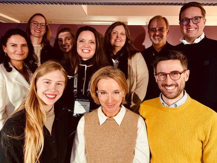 Fotografia de grupo em plano médio, reunindo dez lideranças do setor de turismo em um ambiente interno em Davos. No centro da imagem, Jaqueline Gil sorri abertamente, vestindo uma peça preta com gola branca e ostentando seu crachá de identificação. Ela está cercada por executivos globais, incluindo Robert Brown, Kees Jan Boonen, Lucas Bobe e Jane McFadzean, além de outros participantes do encontro. O grupo está posicionado de forma próxima, todos sorrindo para a câmera em um ambiente com iluminação quente e fundo em tons de bordô e bege. A imagem registra os bastidores do debate sobre os rumos do turismo sustentável.