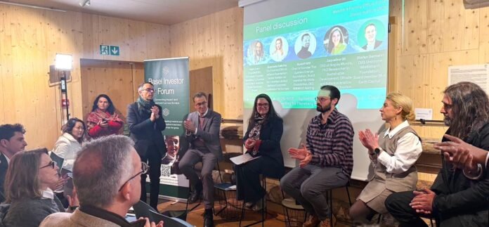 Fotografia de um painel de discussão durante o Wealth and Family Offices Forum Davos 2026. A imagem captura Jaqueline Gil sentada em um banquinho de madeira, participando ativamente de um debate com outros palestrantes em um ambiente rústico e moderno, com paredes de madeira clara.