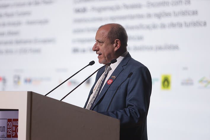 Fotografia de perfil de Toni Sando, presidente da Unedestinos e do Visite São Paulo, capturada enquanto ele fala em um púlpito branco durante um evento. Ele veste um terno azul-marinho, camisa clara e gravata estampada, destacando-se um pin vermelho circular do selo "Made in SP" em sua lapela. Ao fundo, há uma grande tela de projeção com textos e logotipos institucionais desfocados, reforçando o ambiente de uma conferência ou apresentação oficial.